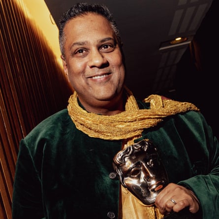 Krishnendu Majumdar – in a scarf, silk tunic and velvet coat – holds an award and smiles next to a wooden wall