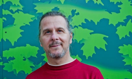 A middle-aged white man with a friendly smile, long gray hair combed back from his face, and a gray goatee, wearing a red long-sleeved shirt and standing outside in front of a what appears to be a mural with a blue background and neon green leaves.