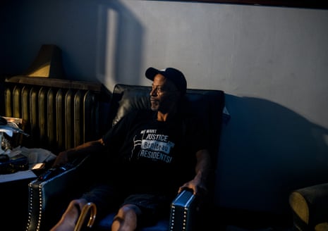 A Black man wearing a black T-shirt and basecall cap sits in a chair on a porch in the dark, looking into what may be the last rays of the sun.