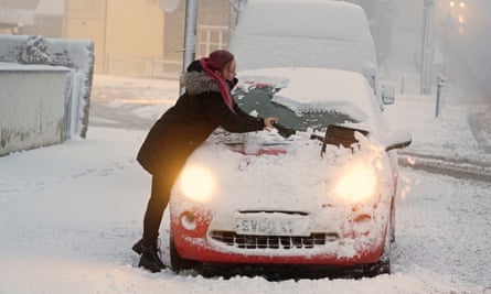 A woman in a heavy coat clears snow from the windscreen of a  small red car on a street with an even covering of snow