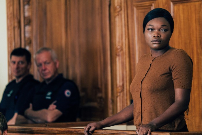 Guslagie Malanda in brown dress stands in the dock in a courtroom. Behind her sit two male police officers