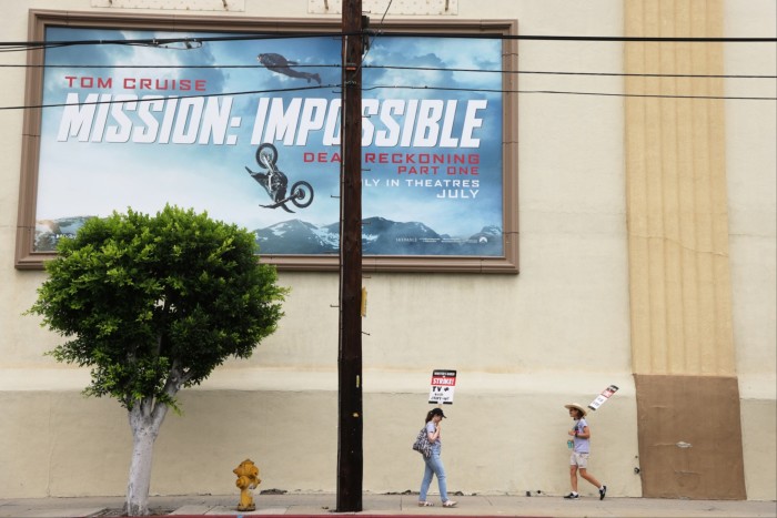 People holding signs walk on a street underneath a billboard advertising Mission: Impossible