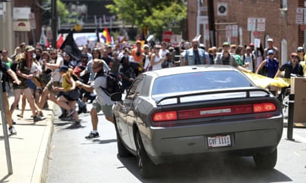 A car drives towards a group of protesters in the sun, with a woman in shorts with a backpack running from the street to the pavement