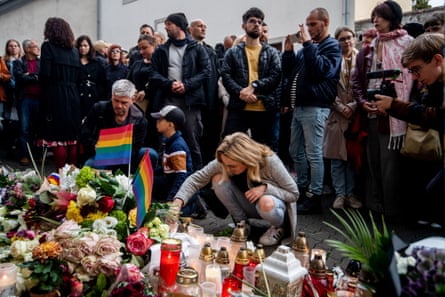 In front of a crowd of people standing, some taking photos, a woman crouches and reaches over to a makeshift memorial of candles and flowers