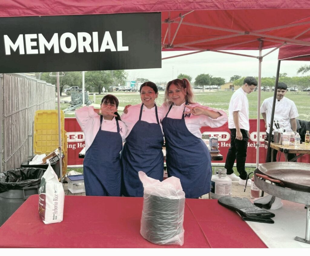Three women in white chef's coats and blue aprons stand behind a table with a red tablecloth. A sign that says Memorial hangs above them and two young men are standing in the background. 