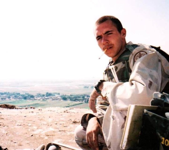 Adam in Iraq, wearing army uniform, crouching, with a cigarette hanging out of his mouth