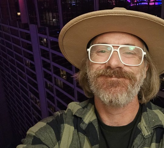 A selfie of Adam with white framed glasses, a beard, long hair and in a wide brim tan hat