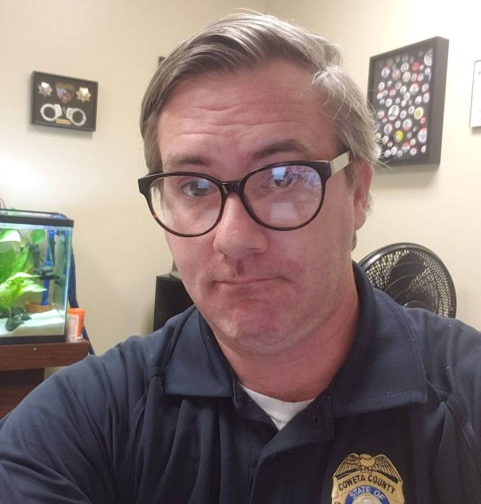 A selfie of Adam in a room with a fish tank, wearing a navy t-shirt with an officer badge