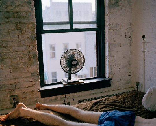 A man lying on a bed in boxers with a fan blowing