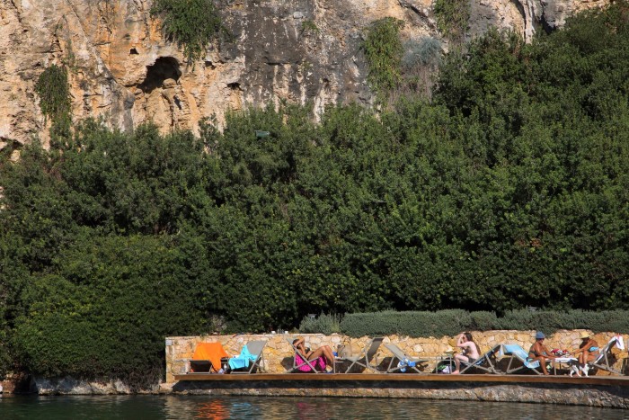 people sunbathing on loungers next to the water against a backdrop of trees and rockface