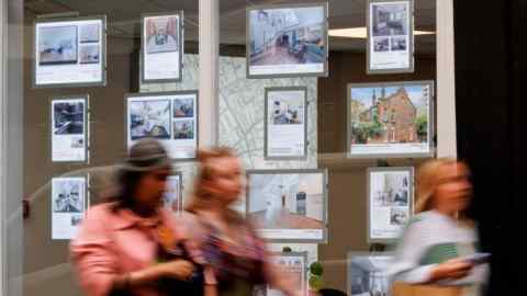 People walk past the window display of an estate agent in London. The display features multiple listings of rental properties with photos and prices.