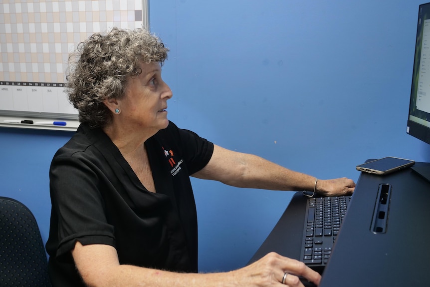 A woman wearing a black shirt working at her computer, against a blue wall.