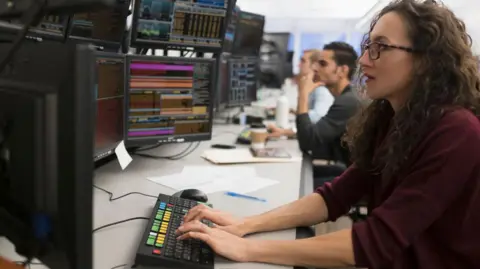 Getty Images Female trader with long hair and glasses sits at keyboard in front of screens