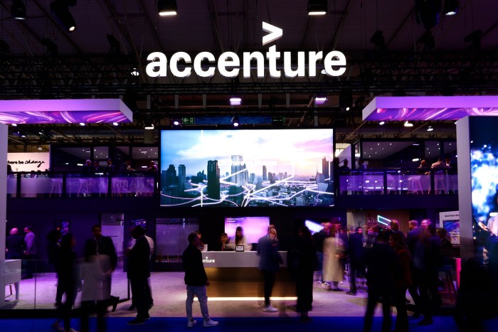 A vibrant Accenture booth at a technology exhibition, featuring a large digital screen displaying a cityscape with illuminated network lines, surrounded by visitors and purple lighting