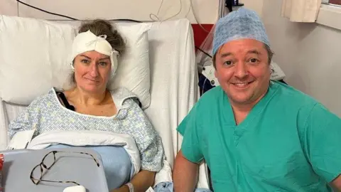 Susannah Morgan Susannah is propped up in a hospital bed with a bandage around her head. She is wearing a hospital gown and her glasses are lying on a table in front of her. Her surgeon, Iain Nixon, is kneeling beside her bed. He is wearing green scrubs and is smiling.