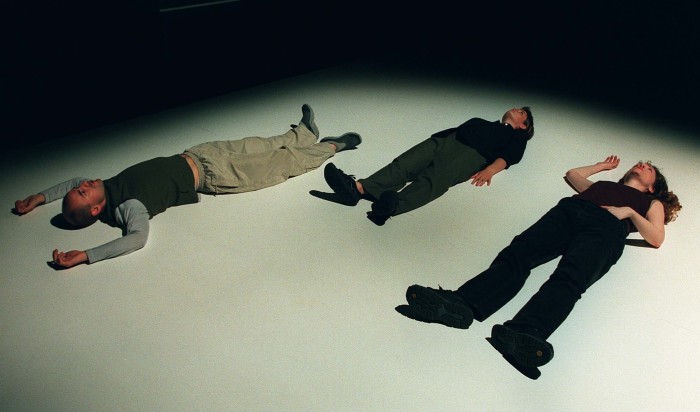Three people, dressed casually, lie prone on a stage. Their eyes are open, and staring upwards