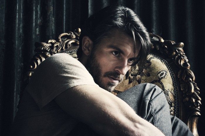 A man with short dark hair and beard sits on an ornate chair with a sultry expression