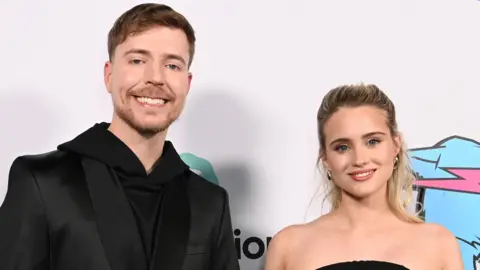 Getty Images MrBeast with Thea Booysen at the premiere of Beast Games in December. MrBeast wears a black suit jacket over a black hoodie and smiles at the camera. He has a short brown beard and short brown hair. Thea wears a sleeveless black dress and has her long blonde hair styled half-up-half-down. 