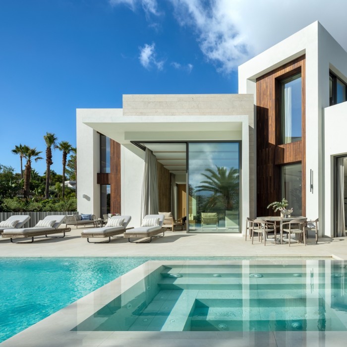 An angular whitewashed modern villa with full height, black-framed windows and a pool with different levels visible under the water