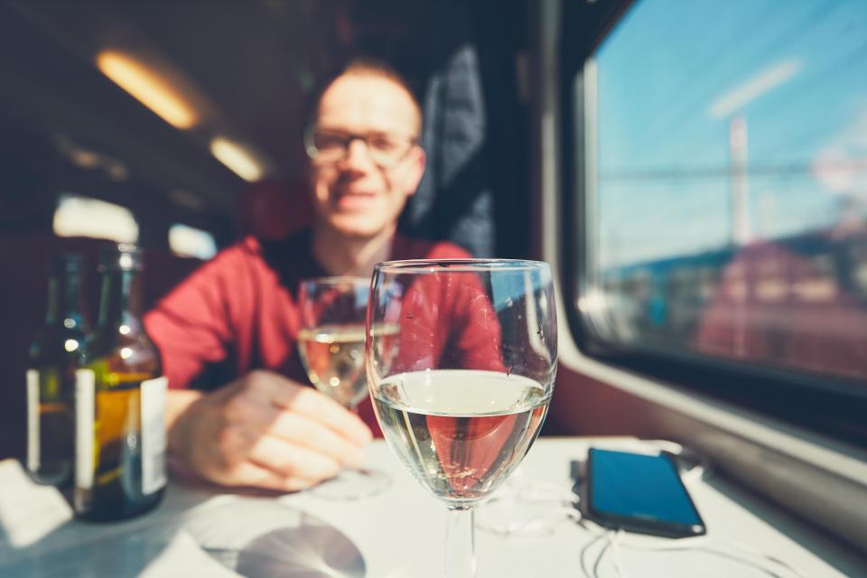 MB087E Rail transportation in sunny day. Smiling young man drinking wine wine on passenger train. Selective focus on the wineglass.