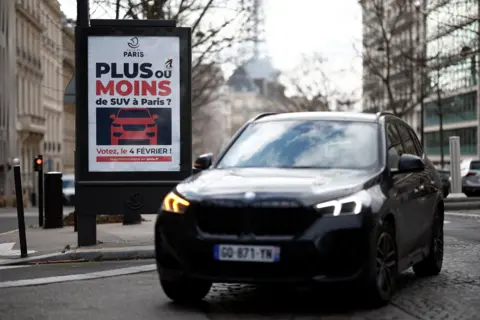 Reuters An SUV drives past a poster in Paris publicising a vote in 2024 over raising parking fees to reduce emissions and increase pedestrian safety