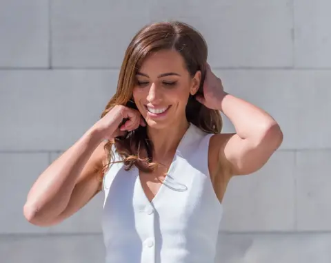 Emma Lynch/BBC Lilly Sabri poses for a photograph. She has long brown hair and is wearing a white waistcoat. She is smiling and looks down while adjusting her hair outside in the sunshine