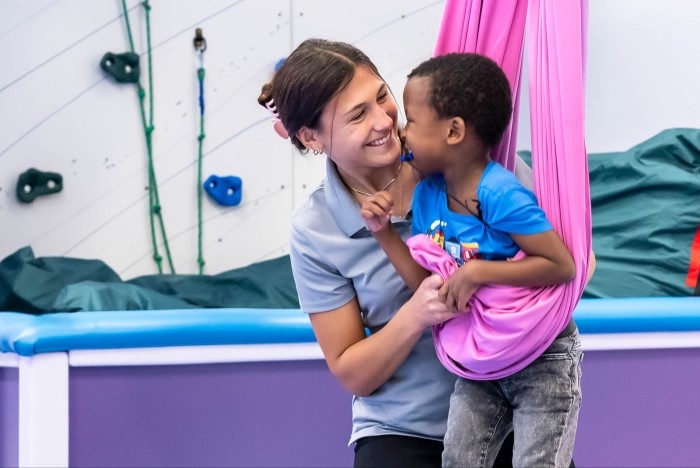 Smiling therapist interacts with a young child in a pink sensory swing at a therapy center
