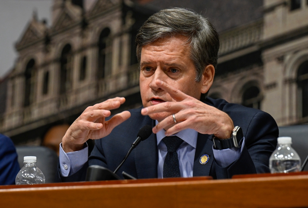 Hoylman-Sigal sitting with hands in front of him at a legislative hearing with a photographic background of the state Capitol.