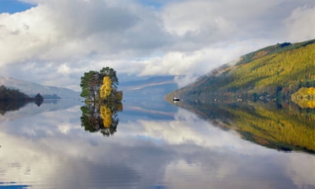 Low-angle view along the mirror-calm water of a loch, with ciouds reflected in the surface and a small island with trees in the middle