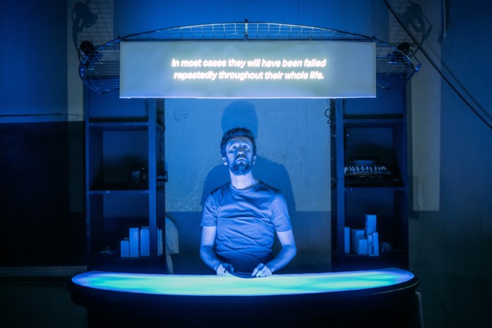 A man stands behind a desk lit by pale blue light