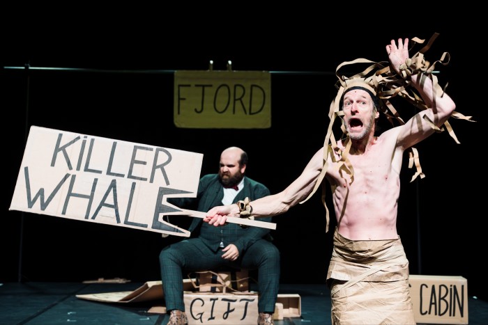 Two performers on stage surroudned by pieces of cardboard adorned with slogans