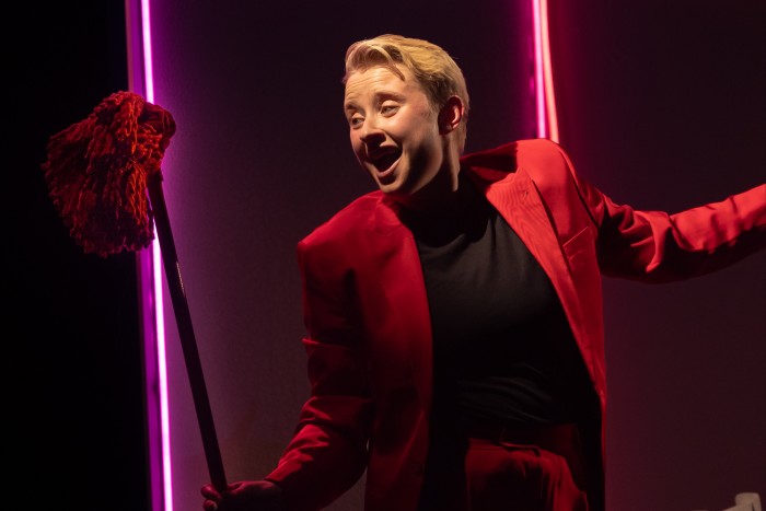 A performer dressed in a suit and backlit by neon lights sings into a mop head