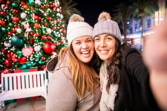 Two friends take a selfie on a phone in front of a Christmas tree.