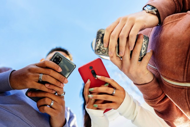 Low angle view of three young people using mobile phones outdoors. Addicted gen z friends holding smartphone outside. 