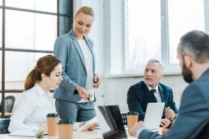 cheerful business coach standing near coworkers and gesturing near digital tablet