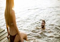 Baden im See. Junges Paar an einem See, während Frau auf Pier sitzt und Mann im See schwimmt.