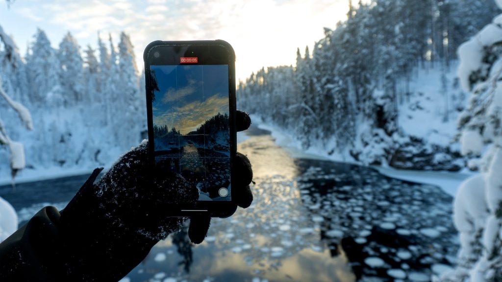 Ruka-Kuusamo: Your Ultimate Guide to Finland’s Natural Wonderland 1 Winter view of a frozen river in Oulanka National Park, Finland, captured through a smartphone held by a gloved hand, with snowy trees and Arctic landscapes in the background.