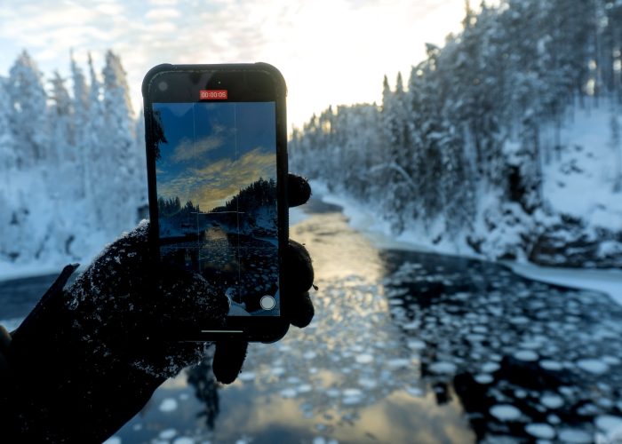 Winter view of a frozen river in Oulanka National Park, Finland, captured through a smartphone held by a gloved hand, with snowy trees and Arctic landscapes in the background.