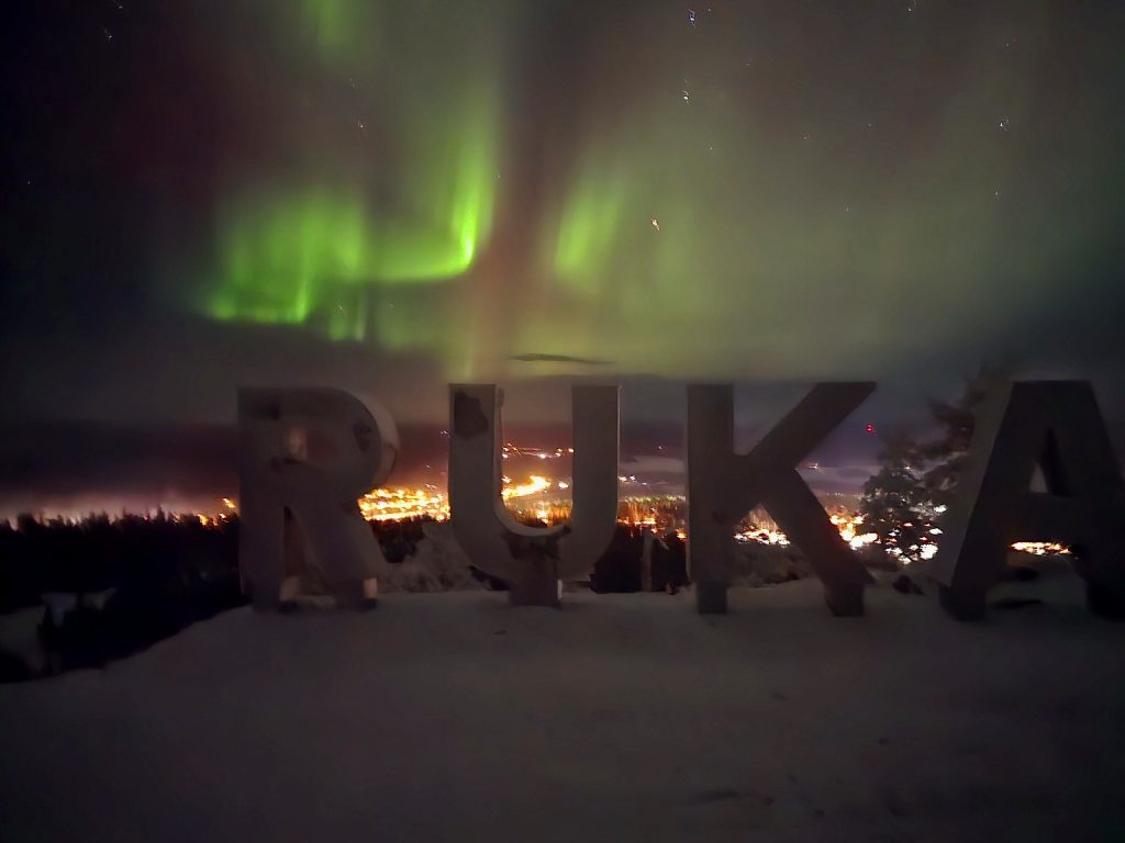 Ruka-Kuusamo: Your Ultimate Guide to Finland’s Natural Wonderland 3 Ruka-Kuusamo sign illuminated by the Northern Lights in Kuusamo, Finland, with a vibrant green aurora dancing in the Arctic night sky above the snow-covered landscape.