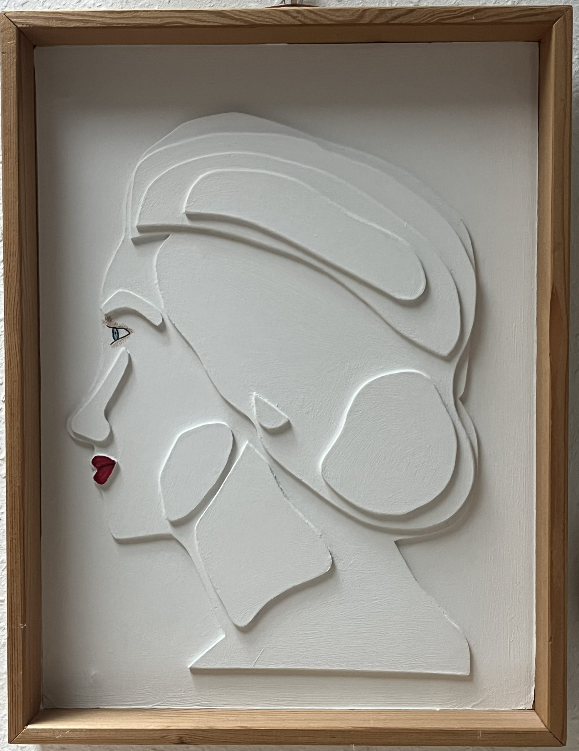Profile of a white bas-relief bust of a woman facing left, with stylized hair, red lips, set in a wooden frame.