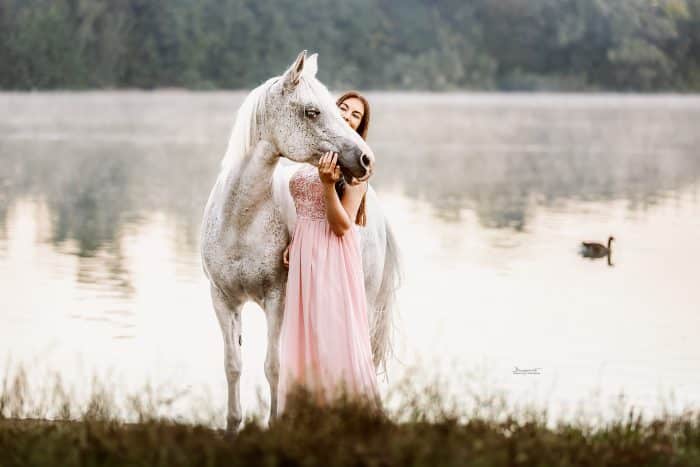 Junge Frau im rosa Kleid steht mit ihrem Pferd am Ufer eines Sees