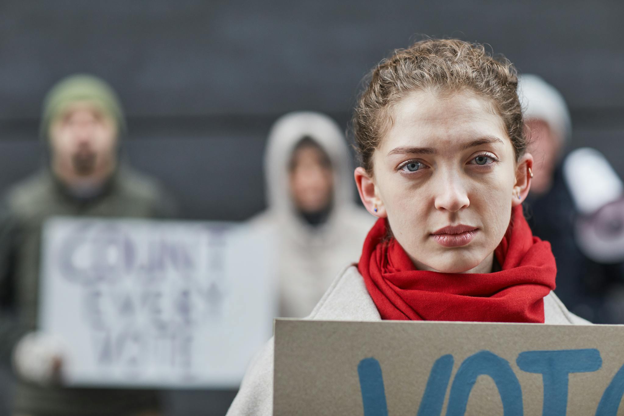 A serious woman in a red scarf at a protest holding a sign, emphasizing importance.