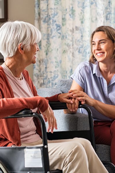 Mature woman comforting senior mom sitting on wheelchair at nursing home. Cheerful woman talking to old disabled mother in wheelchair at elder care centre. Loving caregiver taking care of elderly woman at home.