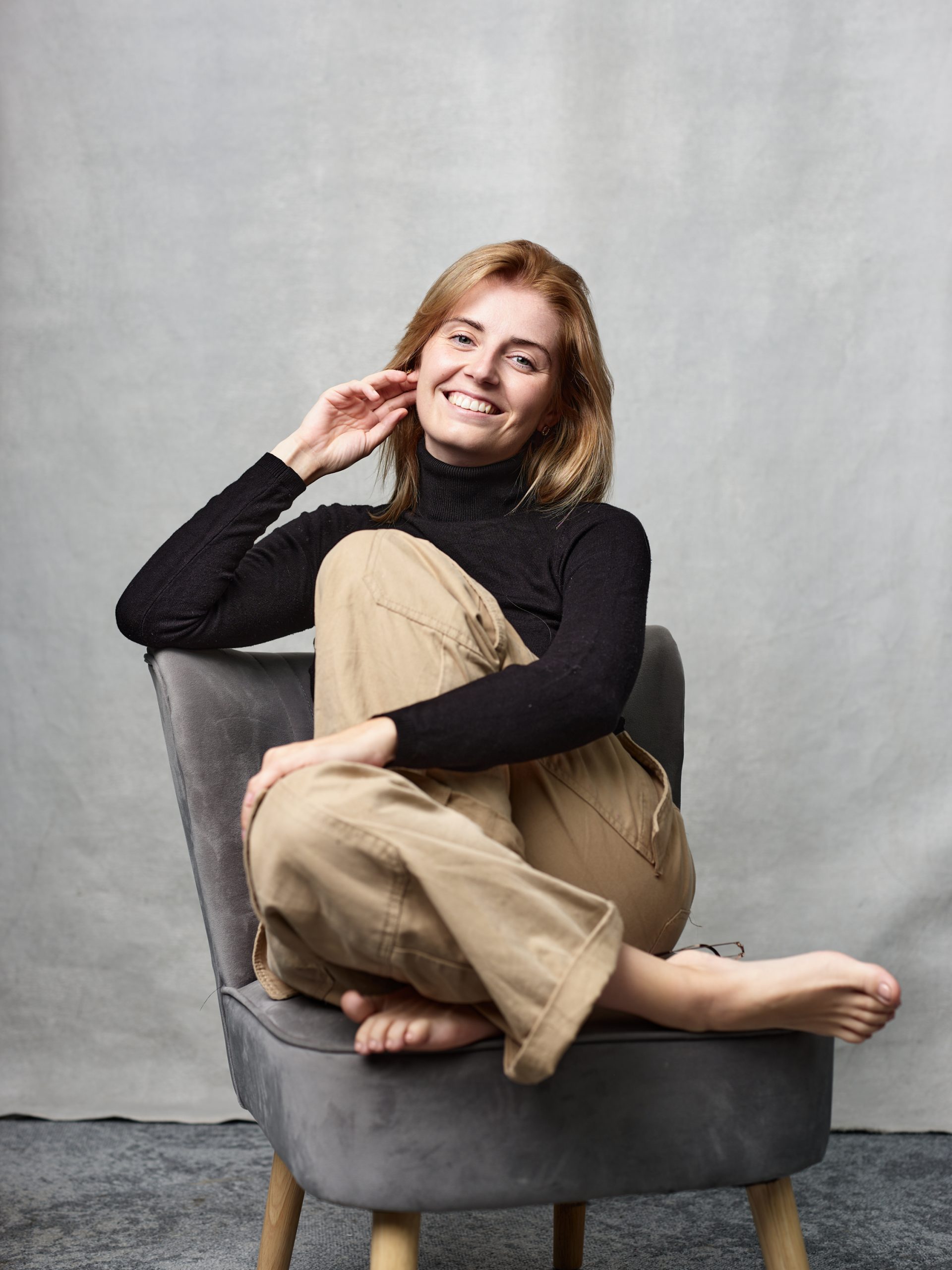 A lady is sat in a grey comfy chair, with her legs crossed, one hand is fiddling with her earring, she has brown hair and is smiling. 