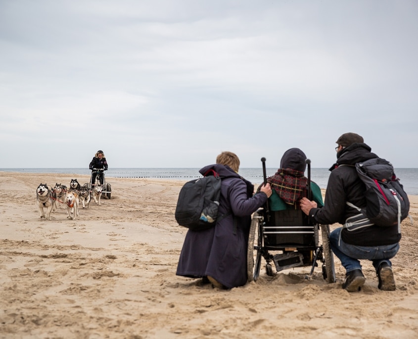 Ein Rollstuhlfahrer steht am Strand mit seinen Begleitern