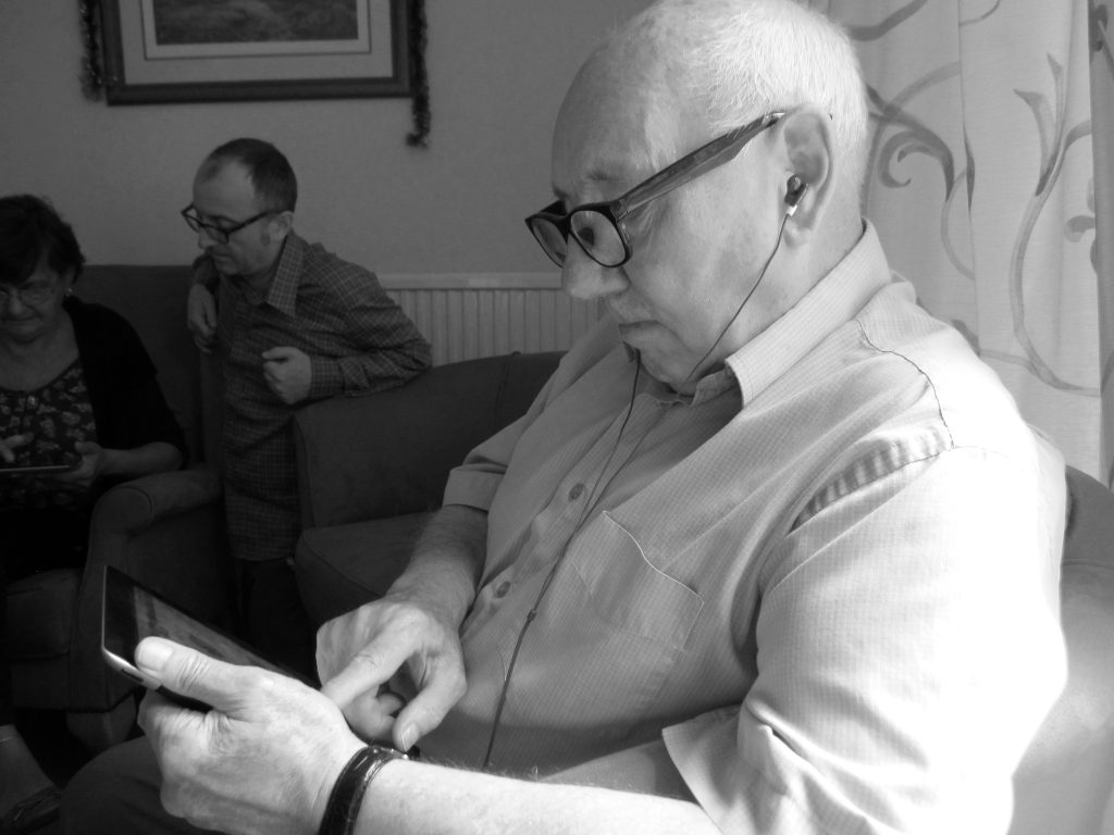 an elderly gentleman wearing glasses and using headphones, using an iPad. In the background Paul is helping a lady.