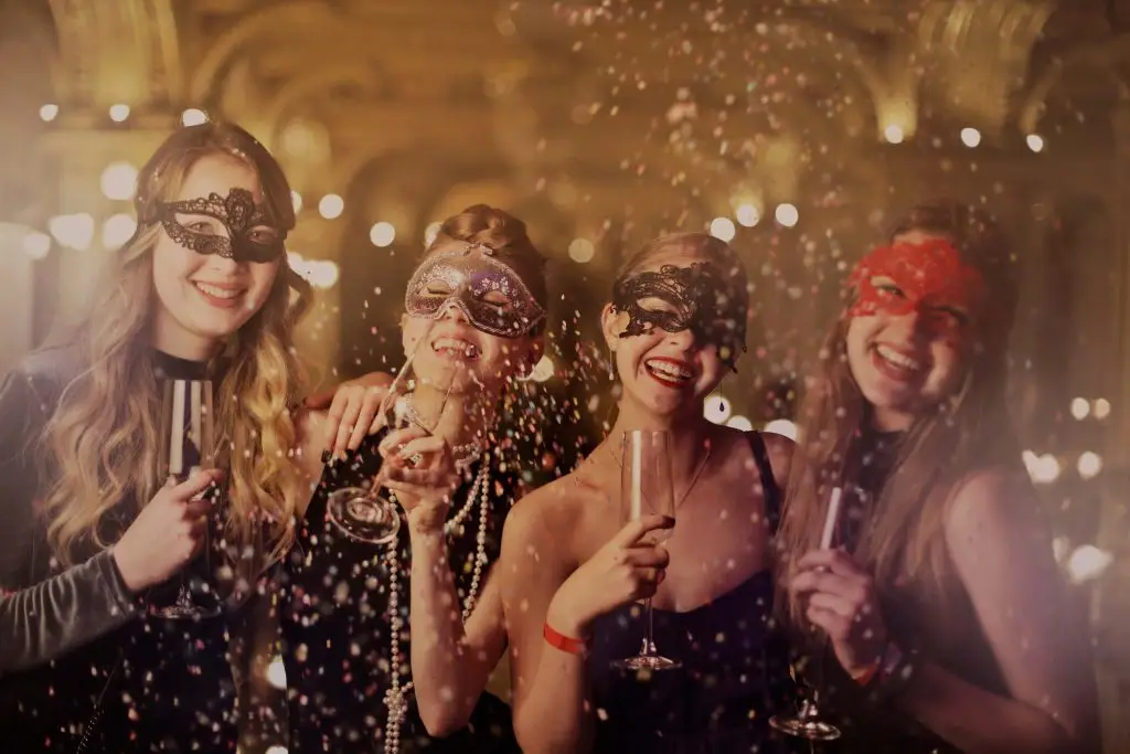 Fire kvinder i masker og festkjoler fejrer til en temafest med champagne og konfetti i en smukt oplyst balsal.