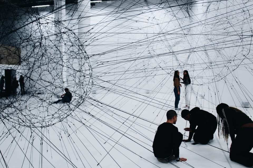 Eine Gruppe Menschen arbeitet gemeinsam an einer küsntlerischen Installation, indem sie schwarze Fäden in einem Raum zu einem Netz verbinden.