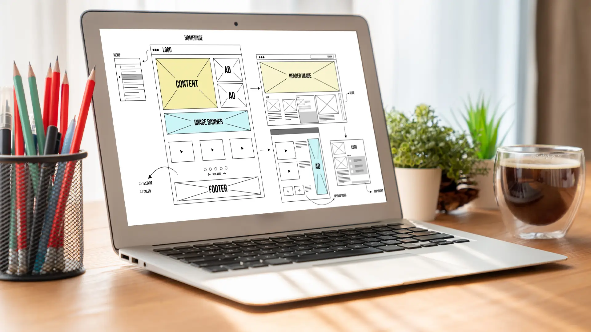 Kunskapsbank 4 Laptop displaying a wireframe layout for a website, placed on a desk with pencils, a coffee cup, and plants in the background.