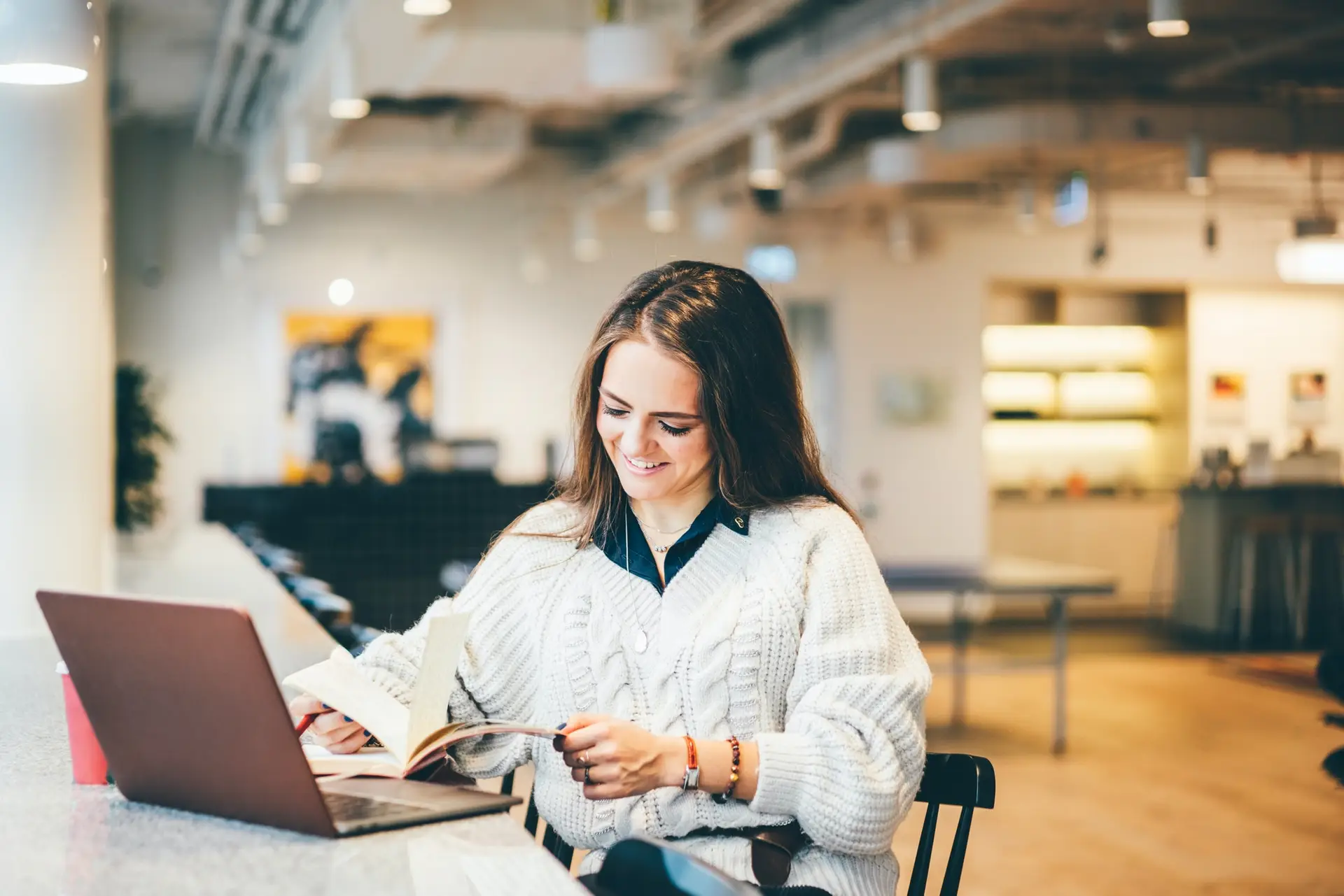 Woman in white sweater reading a book and working on laptop at a modern co-working